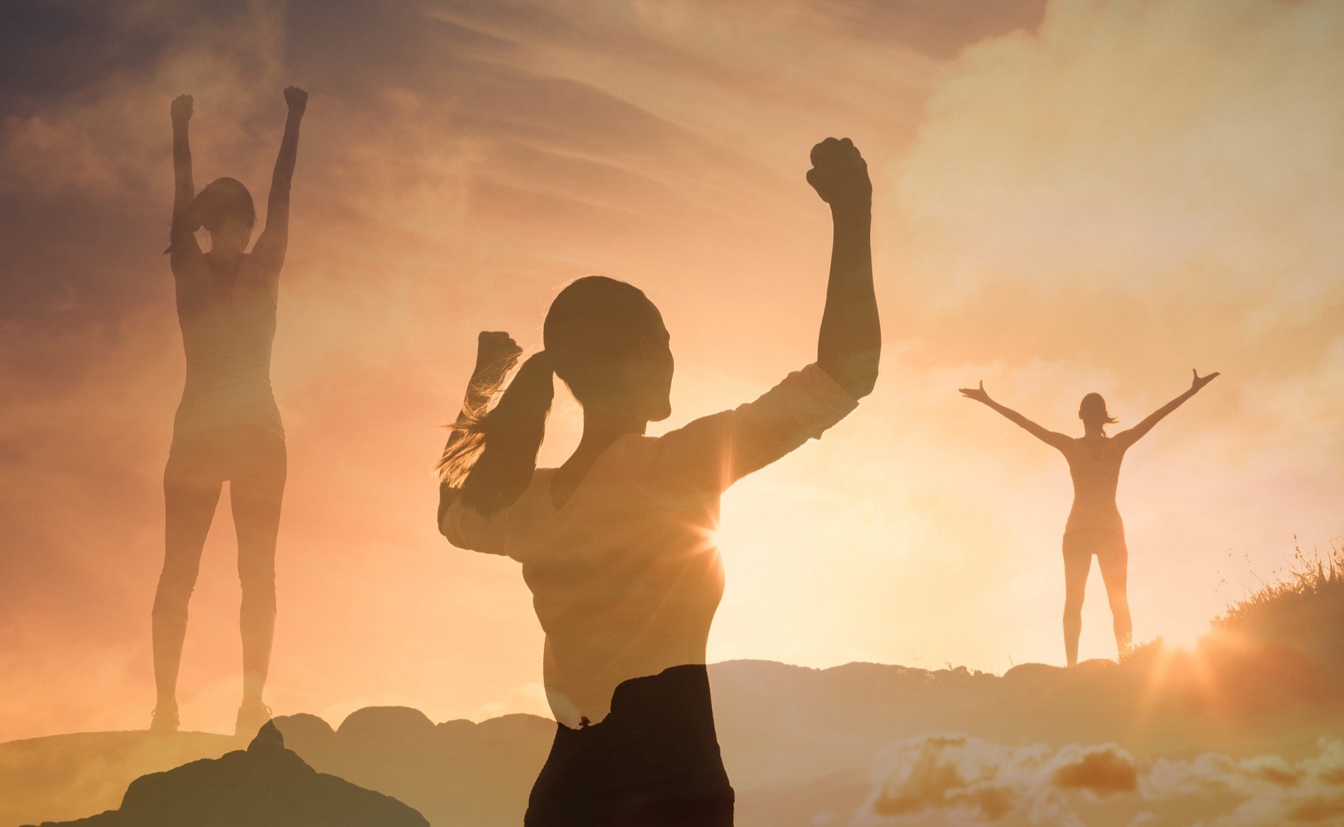 Three people standing on a ridge at sunset with their arms raised in celebration